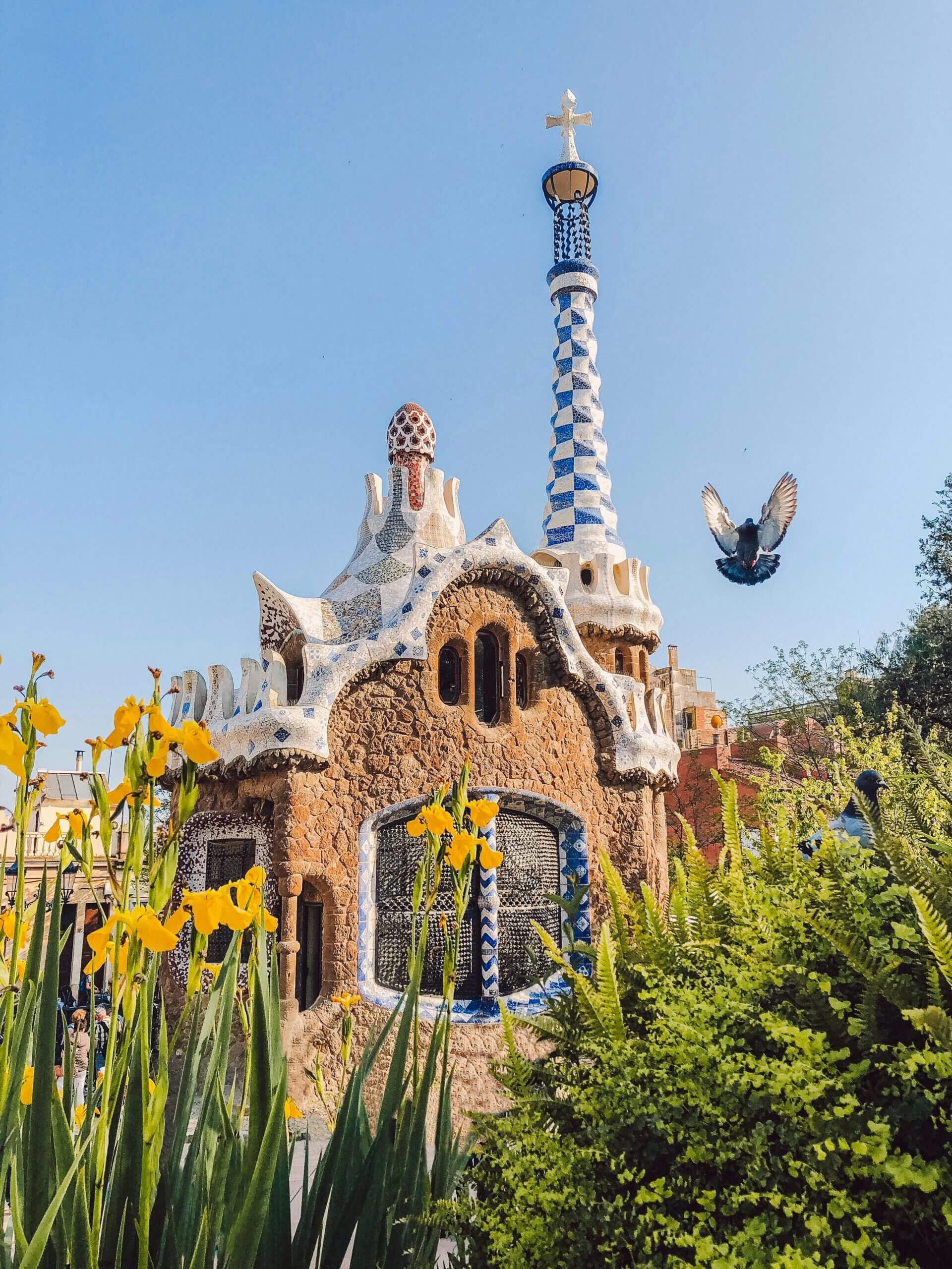 Vibrant image of Park Güell's gingerbread house with blooming flowers and a pigeon flying in Barcelona.